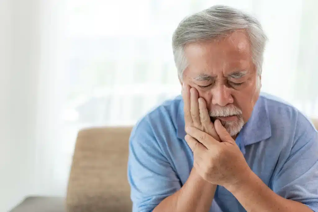 Older man sitting indoors holding his jaw with eyes closed, showing visible discomfort from severe tooth pain or dental issue