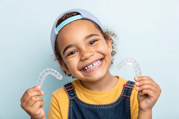 Smiling child wearing a cap and holding two clear aligners, one in each hand.