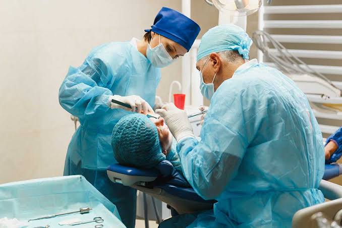 Two dental professionals wearing surgical gowns and masks performing an oral surgery procedure on a patient in a dental chair.